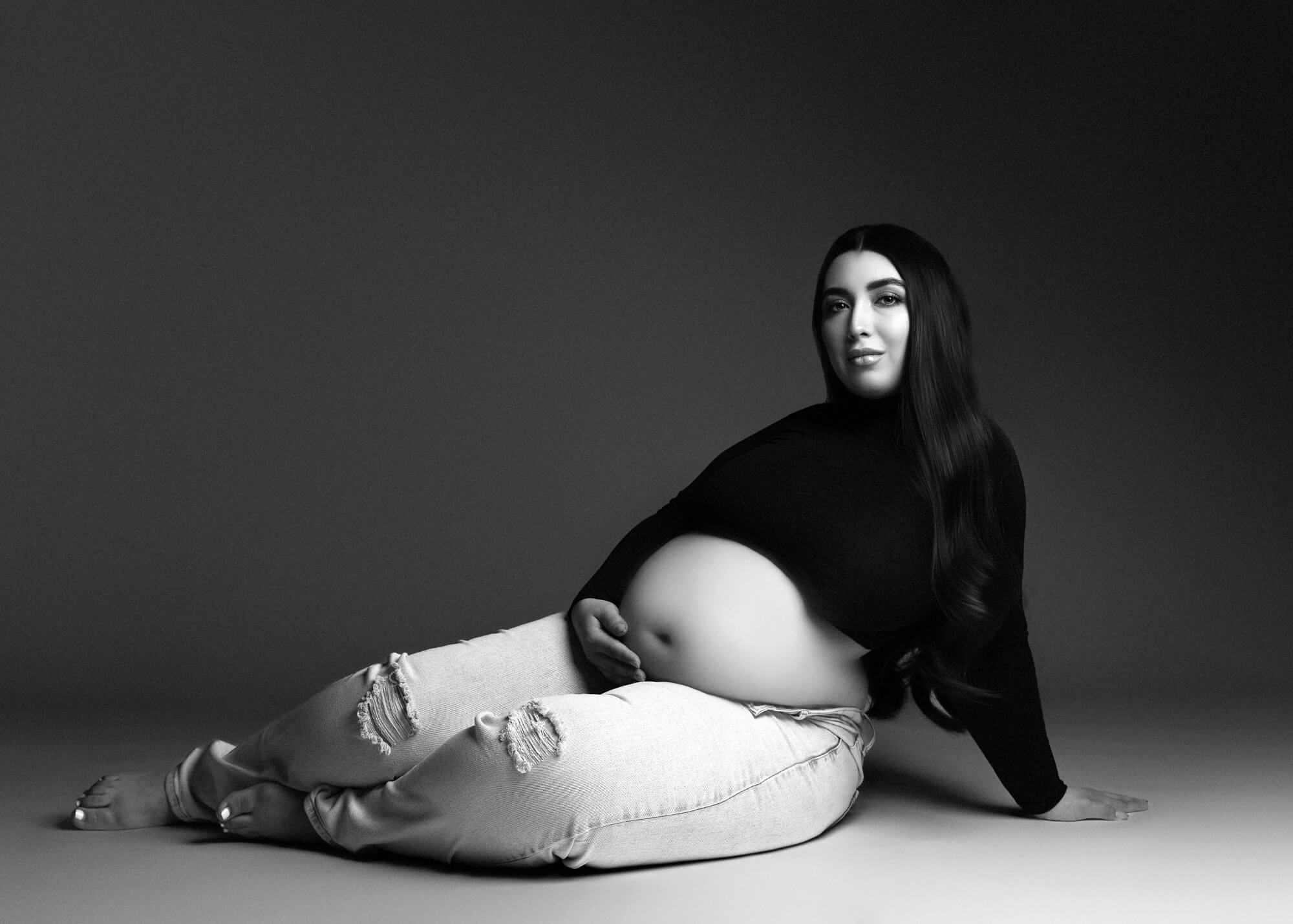 Elegant black-and-white maternity portrait of a woman reclining in jeans and a black turtleneck, photographed in a modern studio setting by Roxy G. Photography.